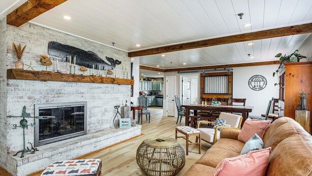 A cozy living room with a fireplace, wooden beams, and a dining area with a table and chairs. - Family Beach Days: Dog-friendly Cape Cod Condo (Dennis Port)