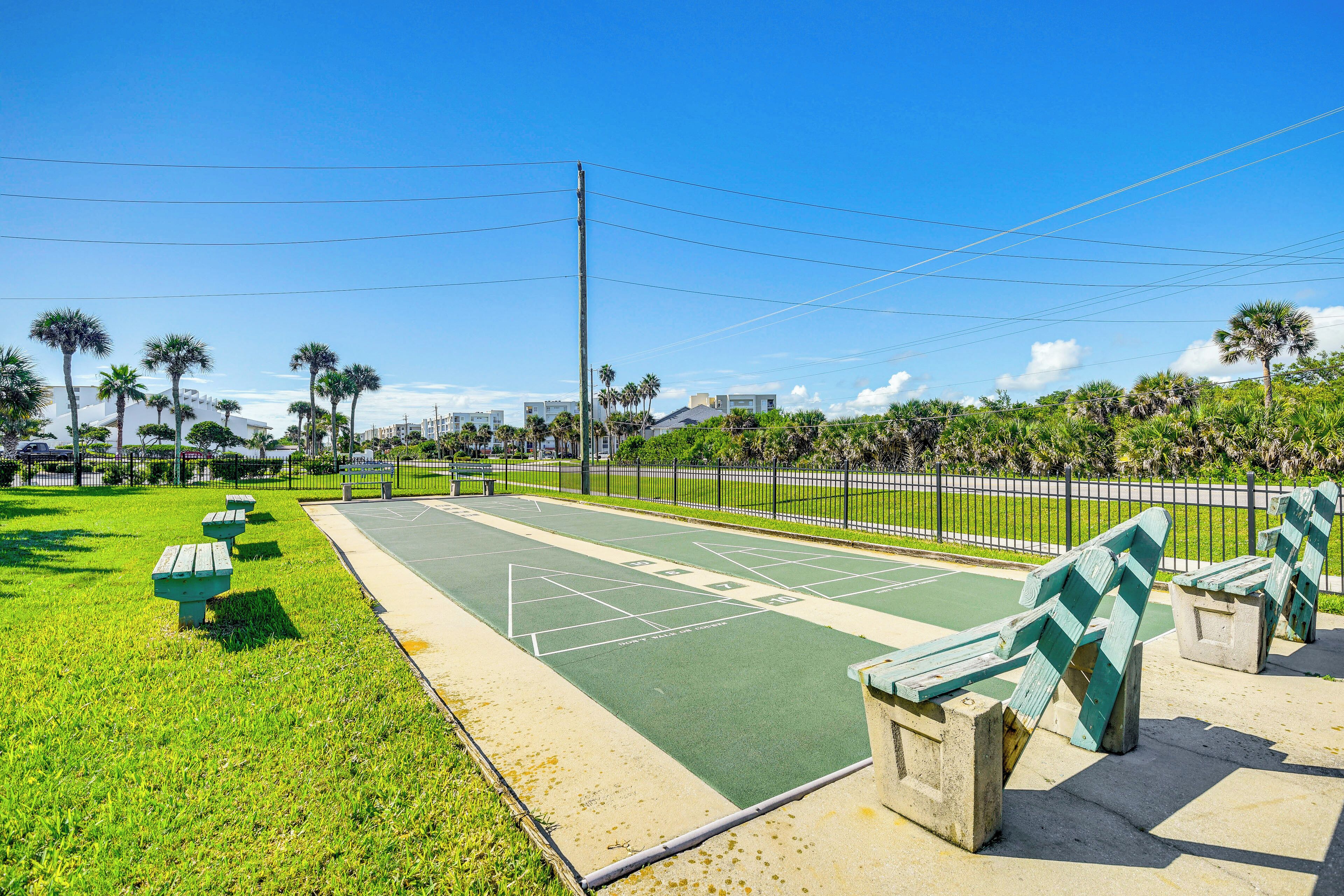A tennis court with green and white markings, surrounded by a fence and palm trees.