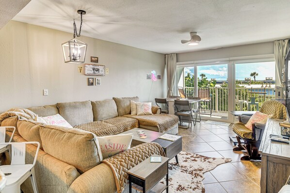 A spacious living room with a large sectional sofa, a dining area, and a balcony with a view. - New Smyrna Beach Condo on Water: Rest, Swim & Play (New Smyrna Beach)