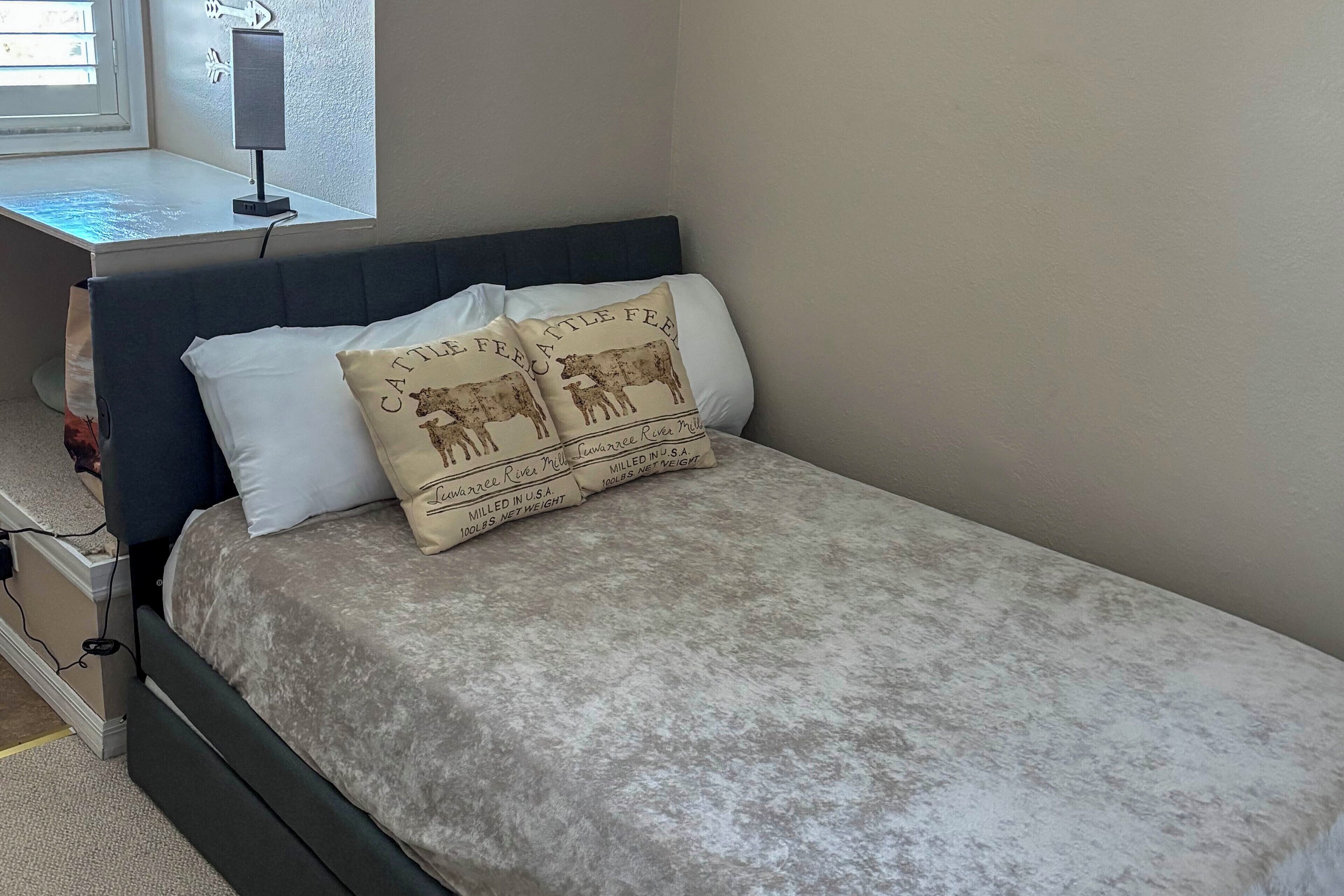 A bed with a grey bedspread and two decorative pillows, a bedside table with a lamp, and a window with blinds.