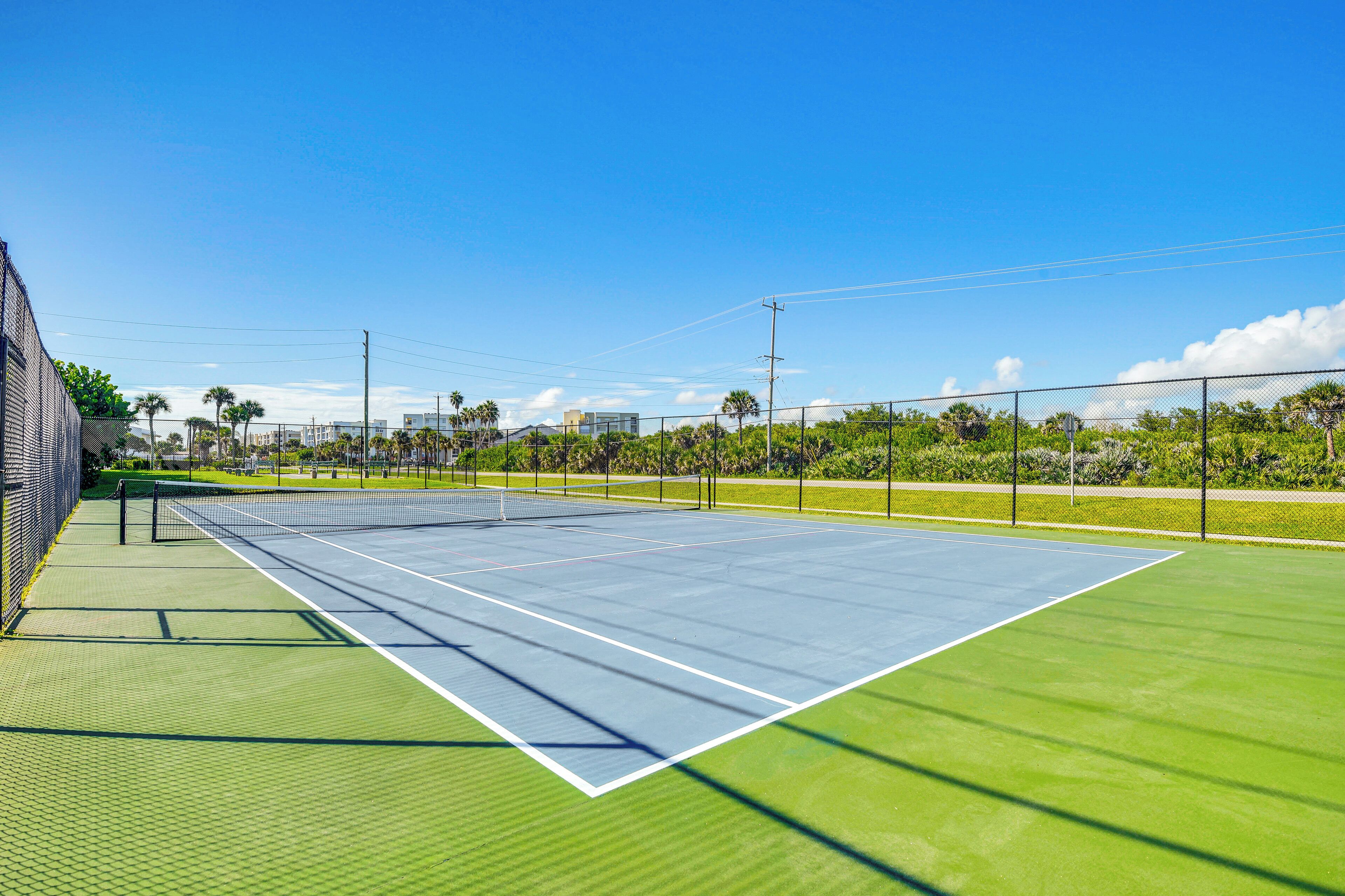 An outdoor tennis court with a net, surrounded by green grass and trees.