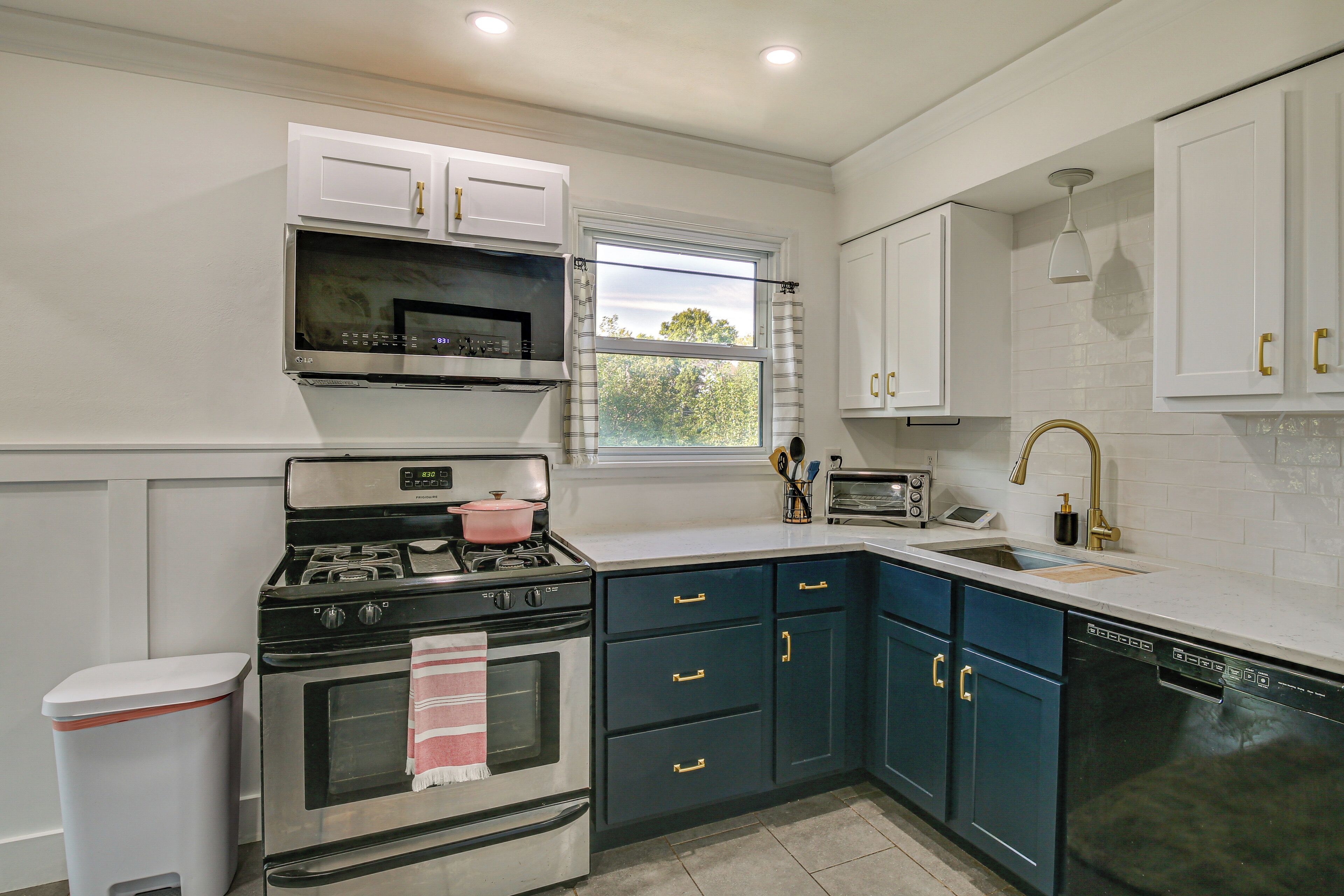 A modern kitchen with white cabinetry, a stainless steel stove, a dishwasher, and a window with a view of trees.