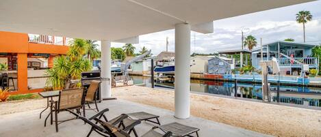 An outdoor patio with lounge chairs and a grill, overlooking a marina with boats and a pool.