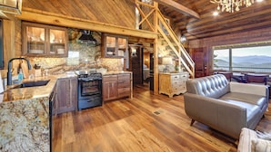 Une cuisine spacieuse avec un plancher en bois, un grand îlot central et un four encastré. Un escalier mène à un niveau supérieur, et un coin salon avec un canapé et une table à manger est également disponible.