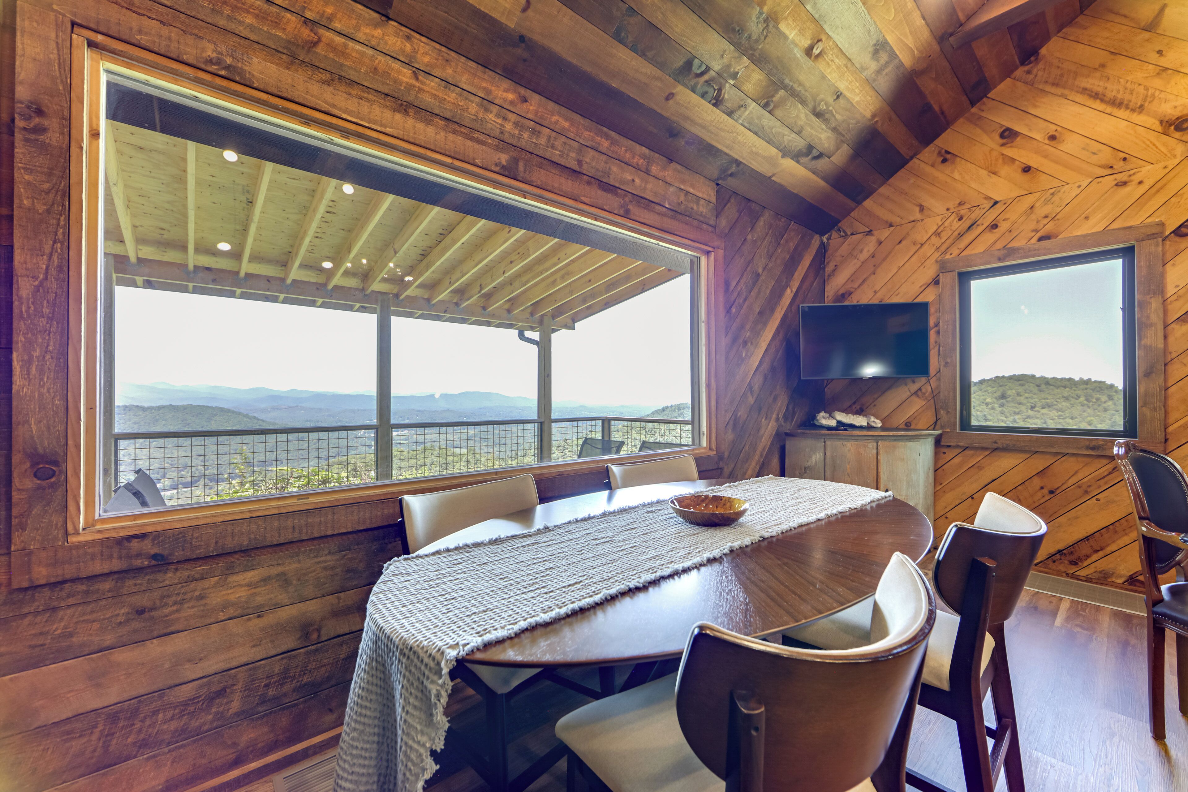 A wooden cabin with a dining table, chairs, a TV, and large windows offering a view of the landscape.