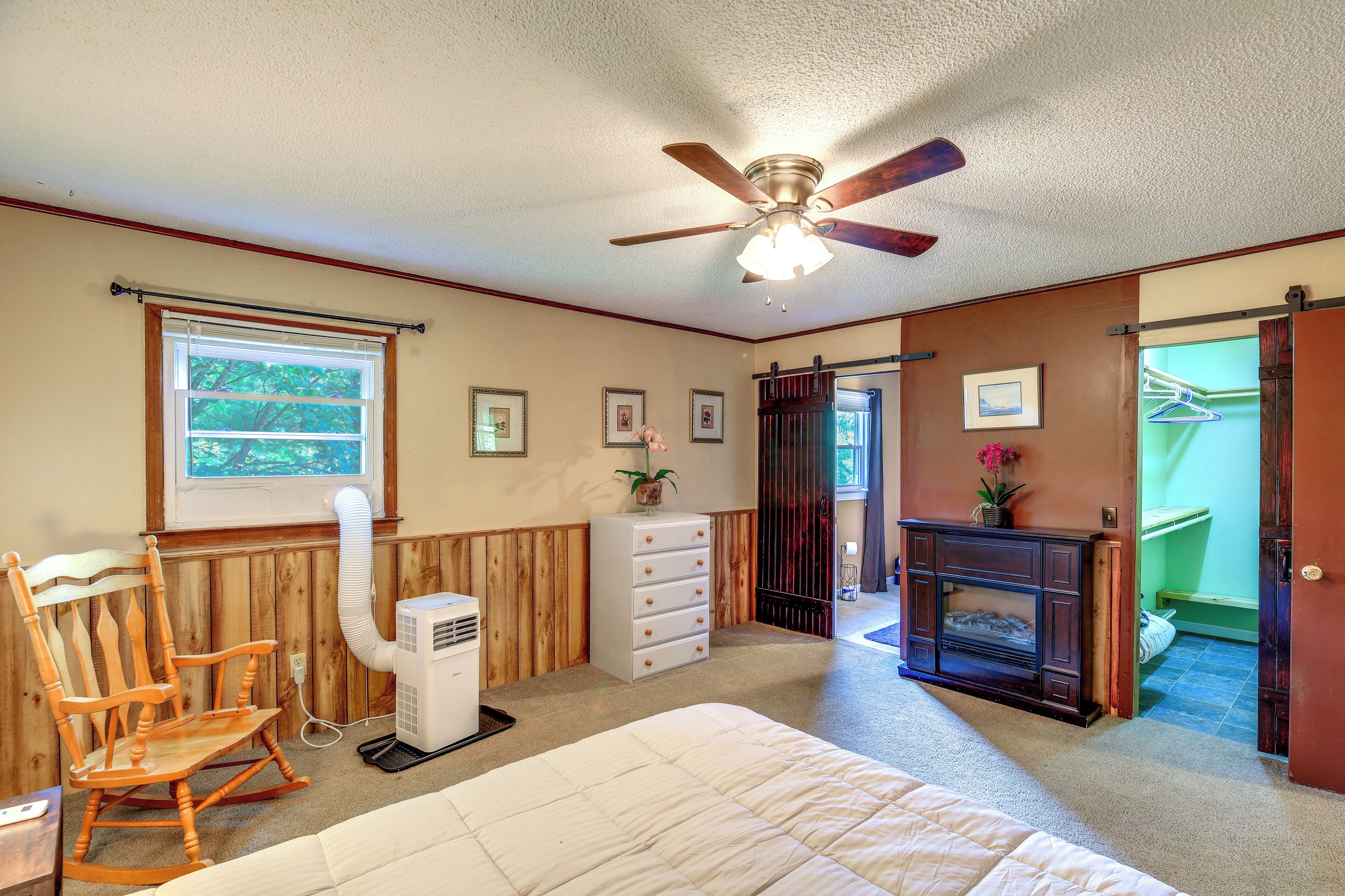 A bedroom with a bed, a rocking chair, a dresser, a heater, a ceiling fan, and a fireplace.