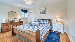 Ein Schlafzimmer mit einem hölzernen Bett, einem Deckenventilator, einem weißen Nachttisch, einem blauen Teppich und einem gerahmten Bild an der Wand.
