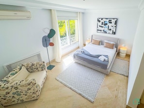 A bedroom with a bed, a chair, a standing fan, a window with curtains, and a wall-mounted air conditioner. - Villa des Artistes in Carthage (Carthage)
