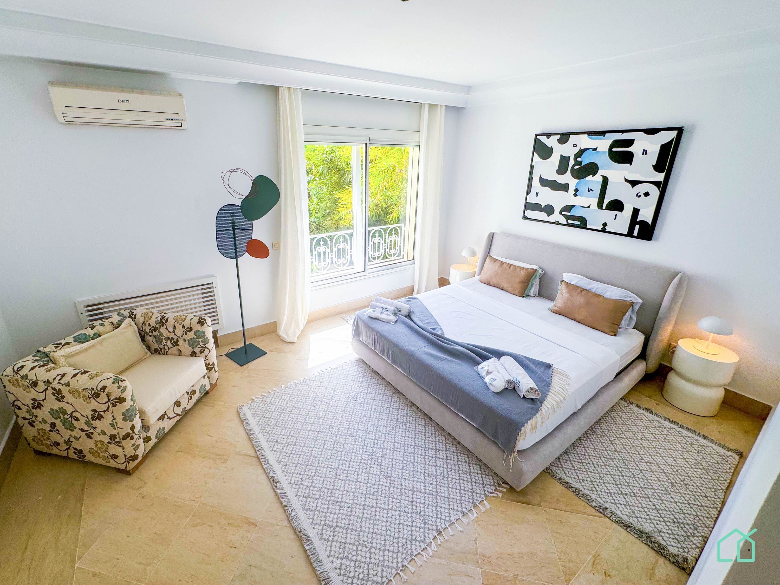 Ein Schlafzimmer mit einem Bett, einem Sessel, einem Standventilator, einem Fenster mit Vorhängen und einem an der Wand hängenden Kunstwerk.