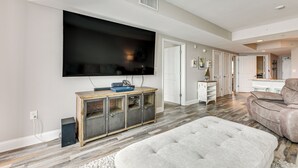 A modern living room with a large flat-screen TV, a wooden cabinet, a plush ottoman, and a beige sofa.