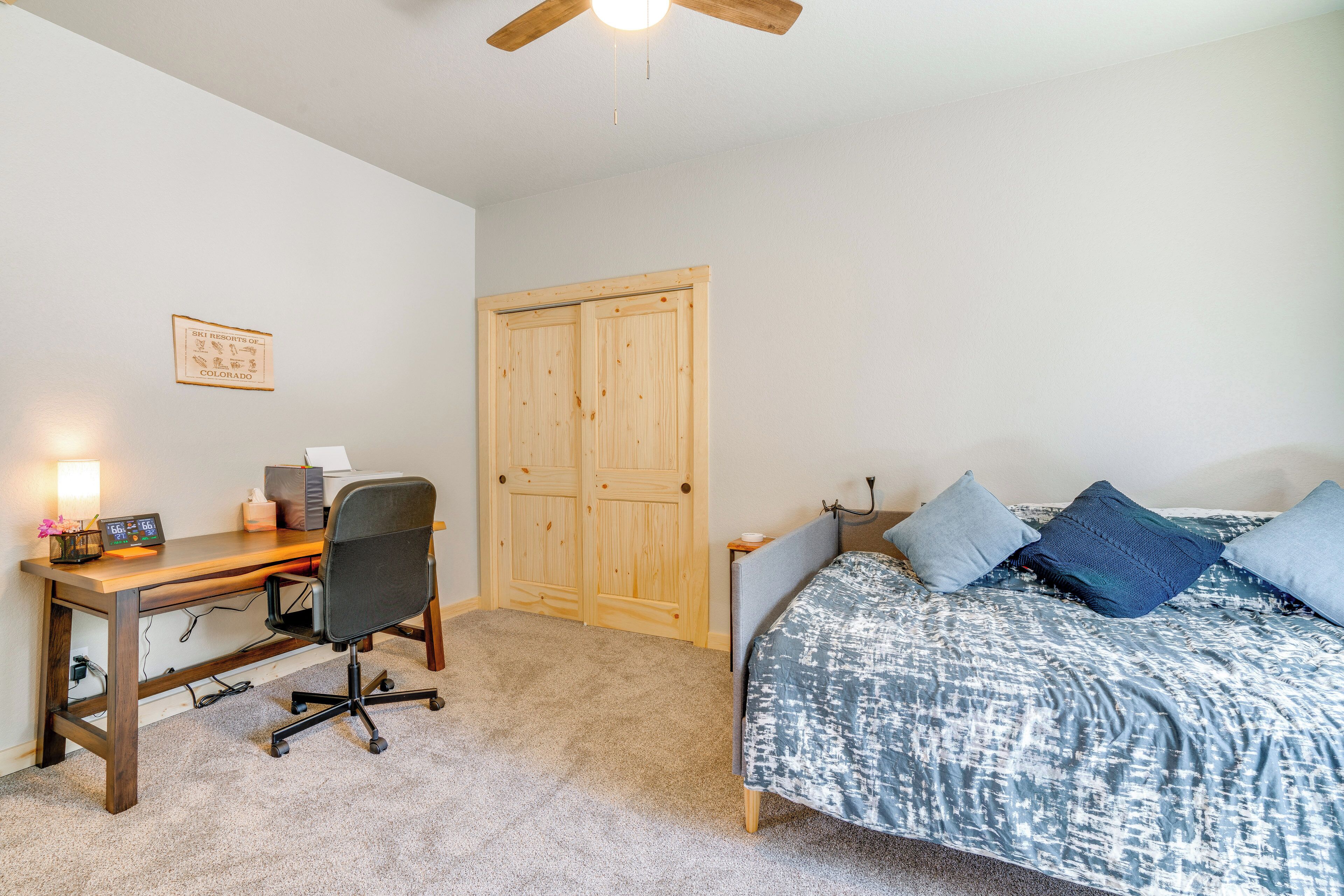 A bedroom with a wooden desk, a chair, a bed with patterned bedding, and a wooden wardrobe.