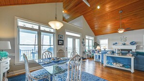 A spacious living area with a dining table, blue patterned chairs, and a blue rug. There are large windows with star decorations, a ceiling fan, and a blue sofa with decorative items. - Riverfront Aurora Home w/ Deck & Private Pier! (Aurora)