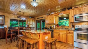 A wooden cabin interior with a kitchen featuring a granite countertop, wooden bar stools, a stainless steel oven, and a large window overlooking greenery. - Appalachian Mtn Cabin Near New River State Park! (Piney Creek)