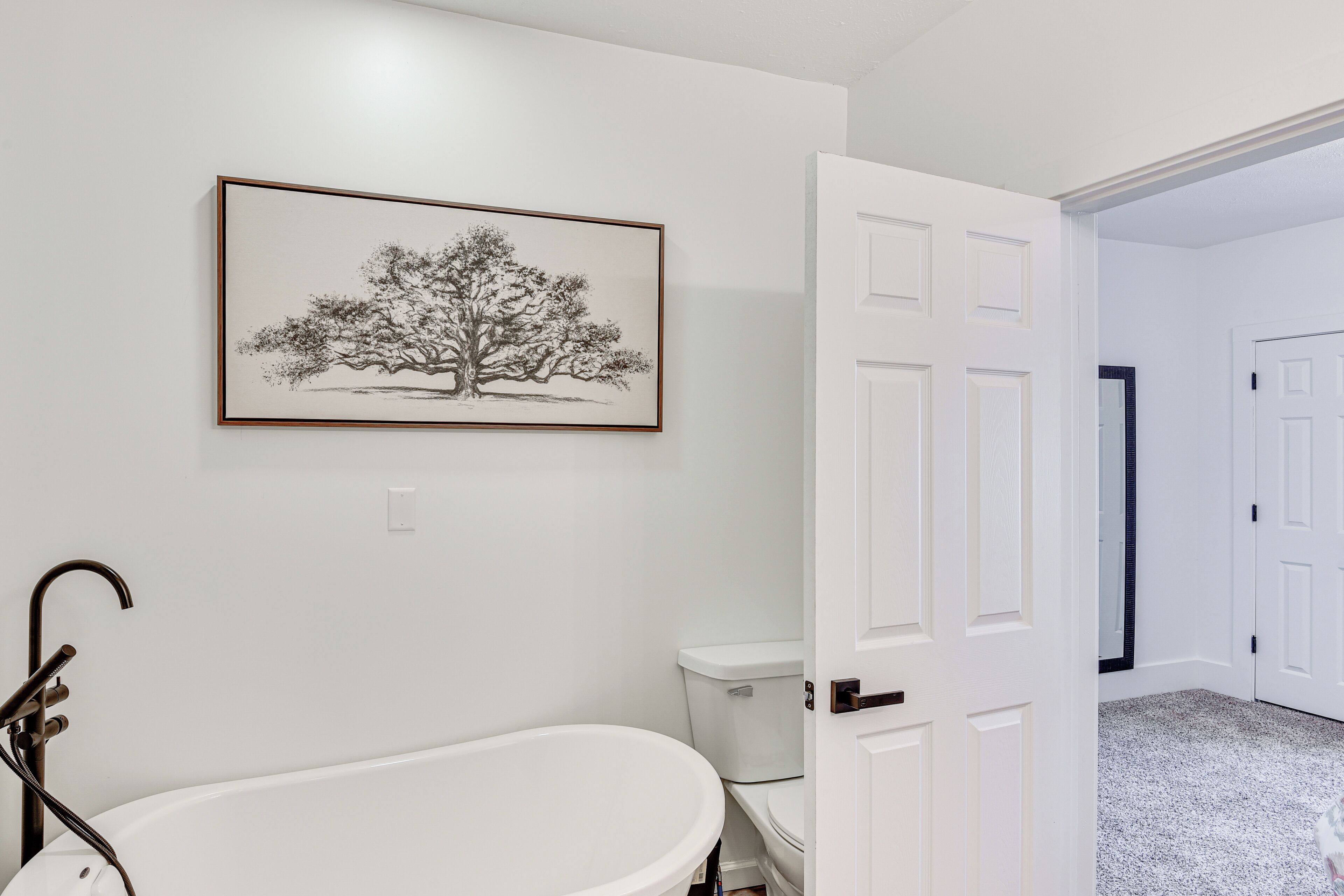 Un baño con una bañera independiente, un inodoro y una obra de arte enmarcadas de un árbol en la pared.