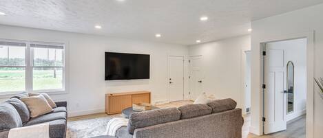 Una sala de estar moderna con un sofá gris, una mesa de centro y un televisor de pantalla plana montado en la pared. Amplias ventanas ofrecen luz natural y vistas al exterior.
