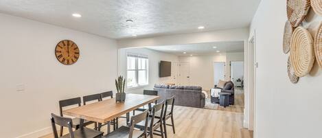A spacious dining area with a wooden table and chairs, a wall clock, and a kitchen in the background.