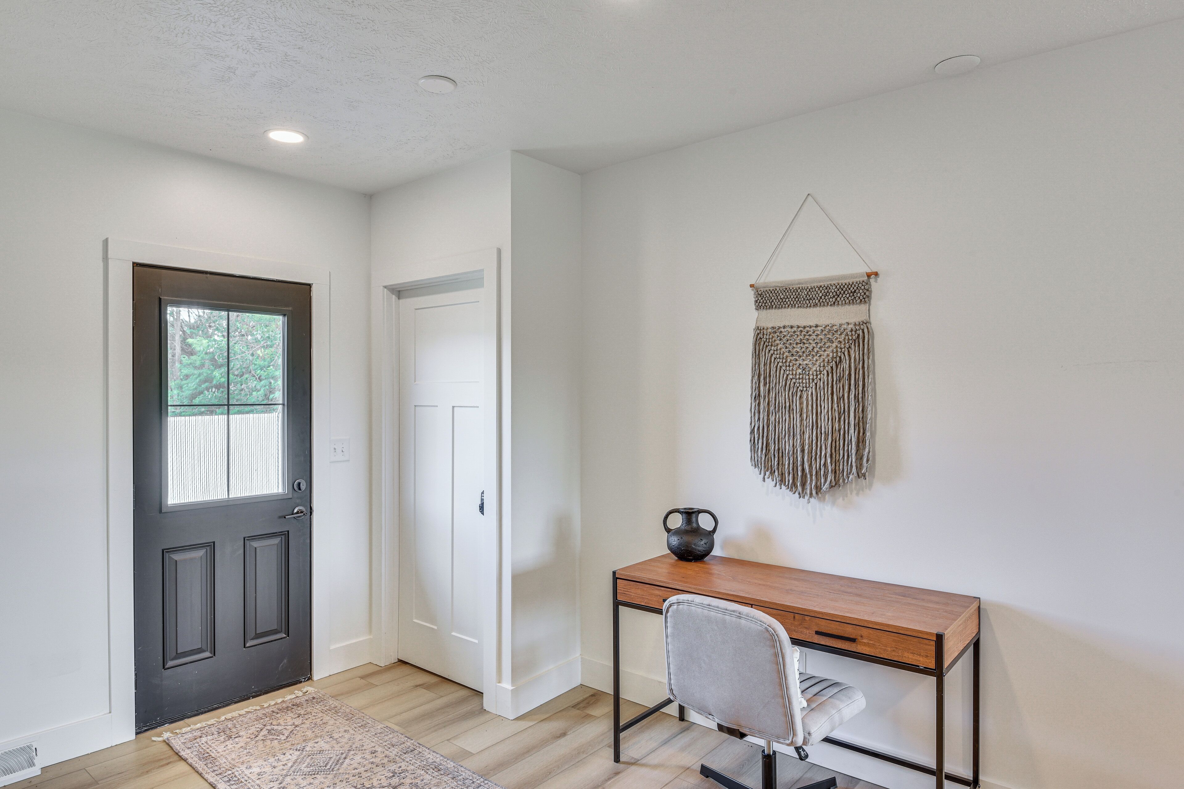 A room with a dark door, a white door, a desk with a chair, and a wall hanging.
