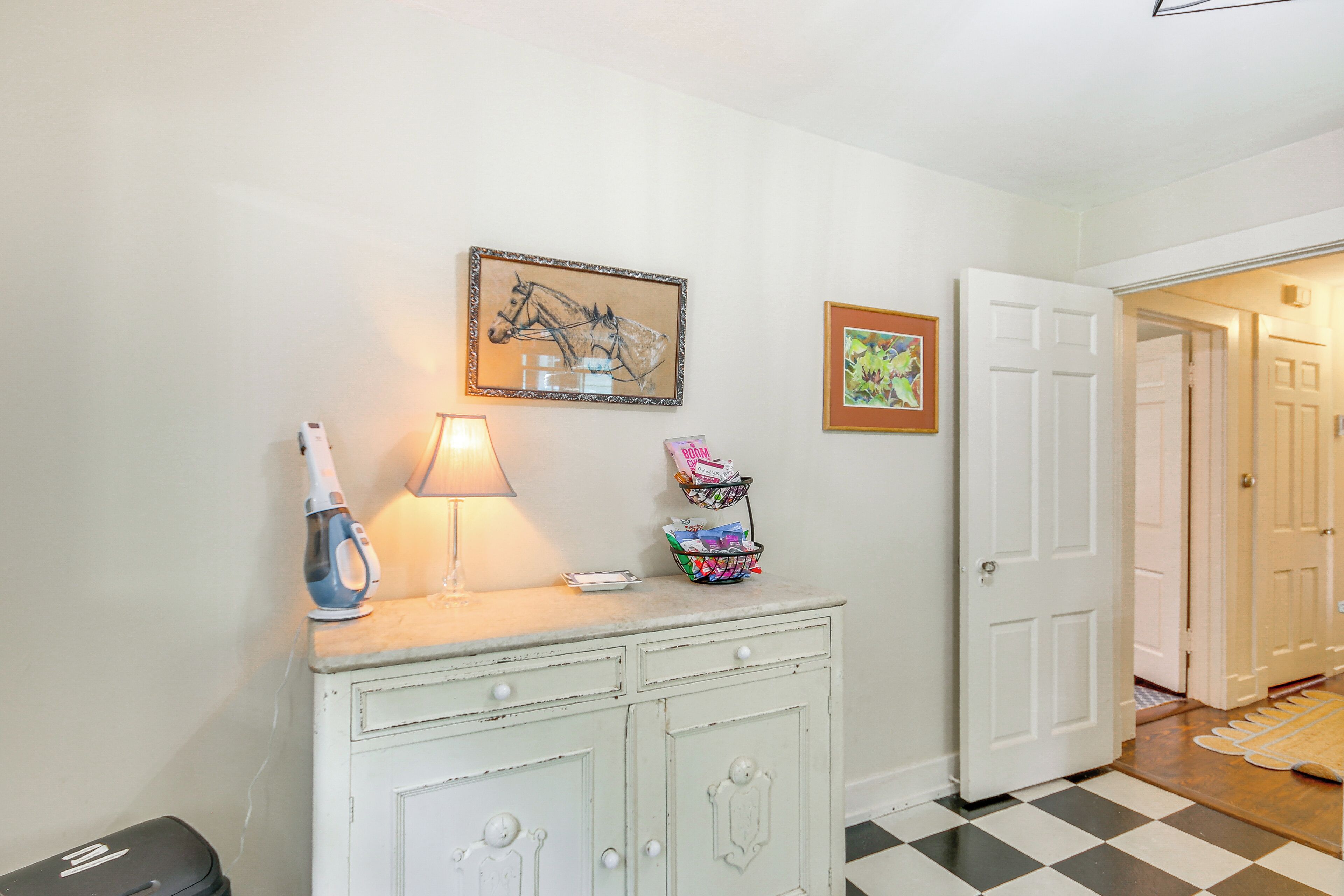 A hallway with a white door, a checkered floor, a white cabinet with drawers, a lamp, and two framed pictures on the wall.