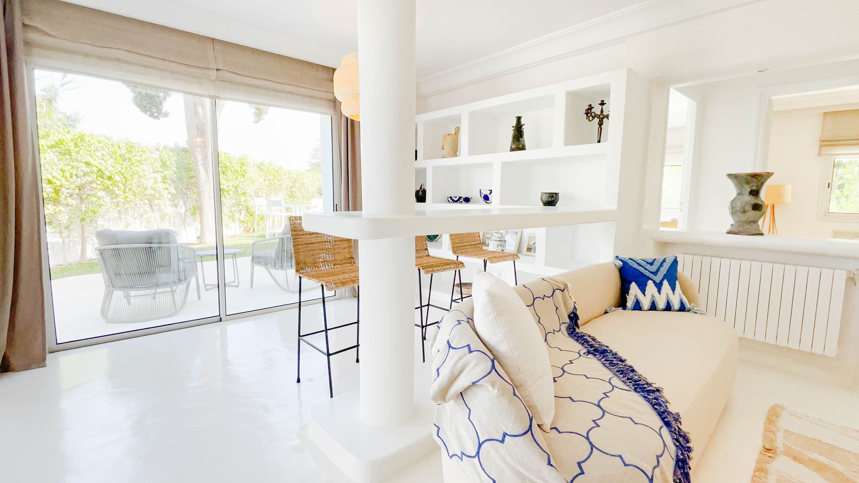 A bright, modern living room with a white sofa, a dining area with wicker chairs, and a view of greenery outside.