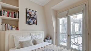 Ein ordentlich bezogenes Bett mit weißem Kopfteil, ein Nachttisch mit Lampe, ein Bücherregal mit Büchern, ein gerahmtes Kunstwerk und eine Balkontür.