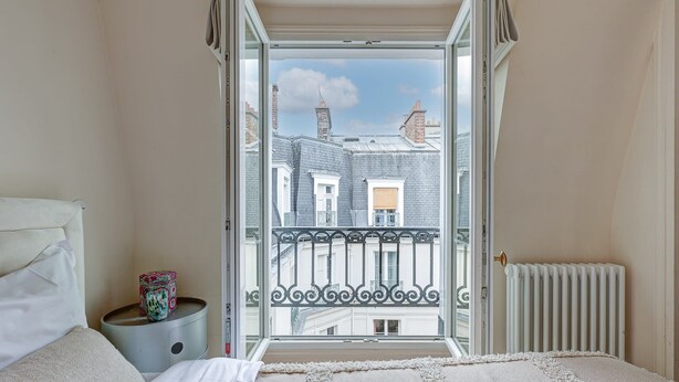 Ein Schlafzimmer mit Bett, Nachttisch, Fenster und Blick auf Gebäude sowie einer Heizung.