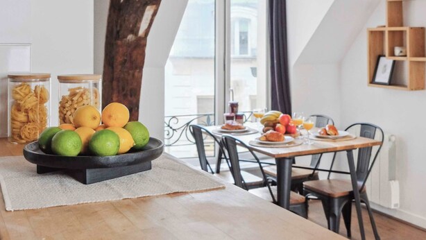 Un área de comedor con una mesa lista para el desayuno, un cuenco de fruta y vistas de la ciudad a través de una ventana.