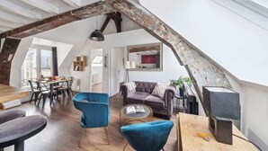 Un espacio habitable estilo loft con vigas de madera expuestas, una zona de comedor con una mesa y sillas de madera, un sofá morado y un ventanal que proporciona luz natural.