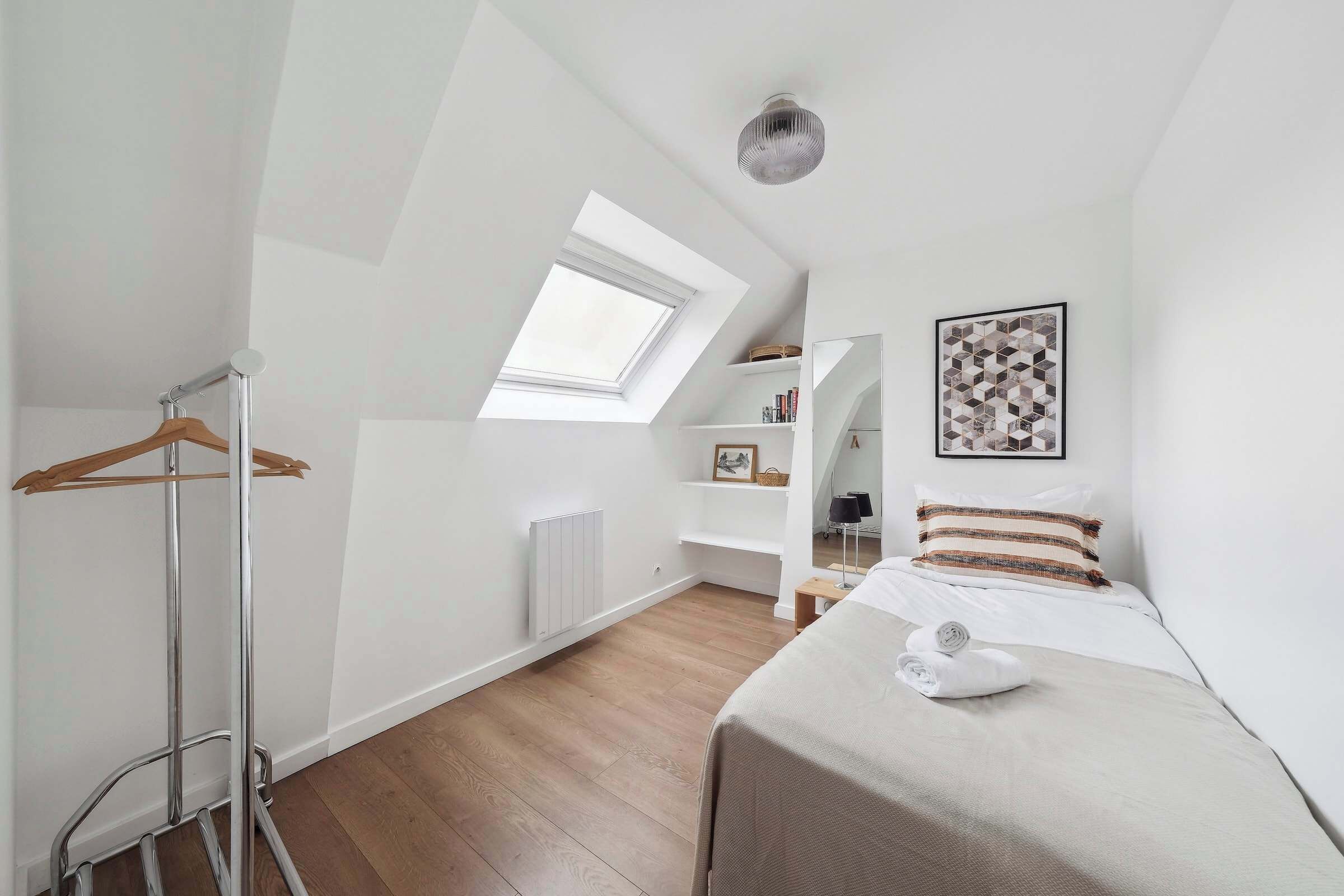 A modern bedroom with a skylight, a bed with a rolled towel, a hanging clothes rack, and a white radiator.