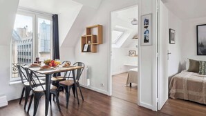 Comedor con mesa lista para cuatro personas, vista de la ciudad a través de un ventanal grande, y un dormitorio tipo loft con cama y vista de la ciudad.