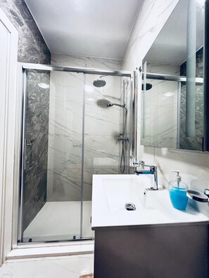 A modern bathroom with a glass-enclosed shower, a white sink with a soap dispenser, and a large mirror.