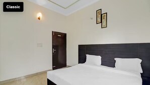 A classic hotel room with a large bed, a door, a wall-mounted light, and framed pictures. - Hotel Olive Tree (Agra)