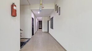 A hallway with a fire extinguisher mounted on the wall, a ceiling light fixture, and framed pictures on the wall. - Hotel Olive Tree (Agra)