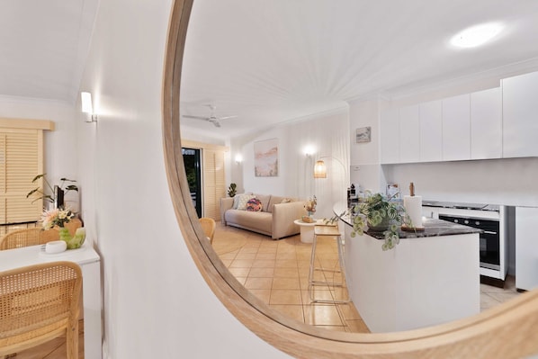 A modern kitchen with white cabinets, a dining area with wicker chairs, and a living room with a beige sofa.