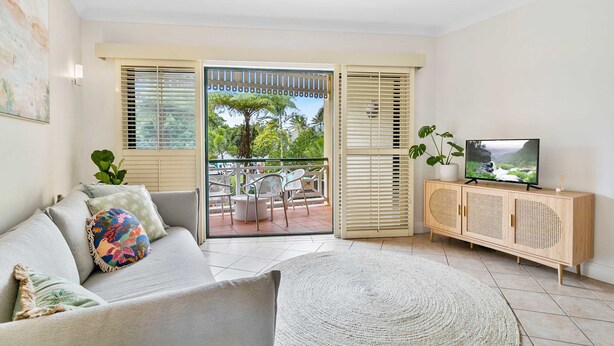 A living room with a sofa, a TV on a wooden cabinet, and a view of a patio with chairs and a table. - Cozy Waterfront - Free Parking Pool (Cairns North)