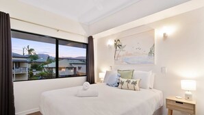 A hotel room with a large bed, a nightstand with a lamp, a painting on the wall, and a view of houses and palm trees outside the window. - Cozy Waterfront - Free Parking Pool (Cairns North)