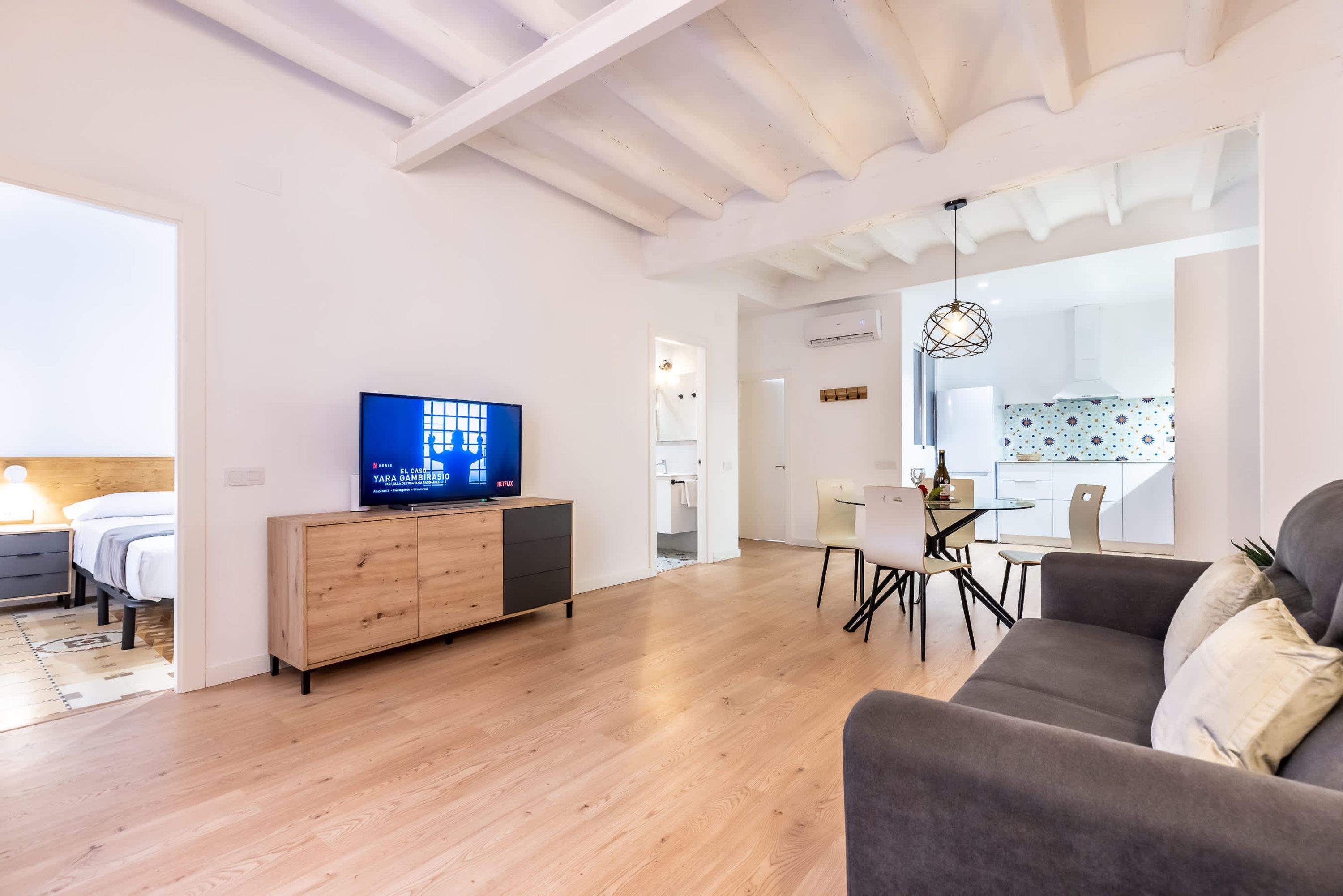 Una sala moderna con un mueble de madera, un televisor de pantalla plana, un sofá gris y un comedor con mesa y sillas.