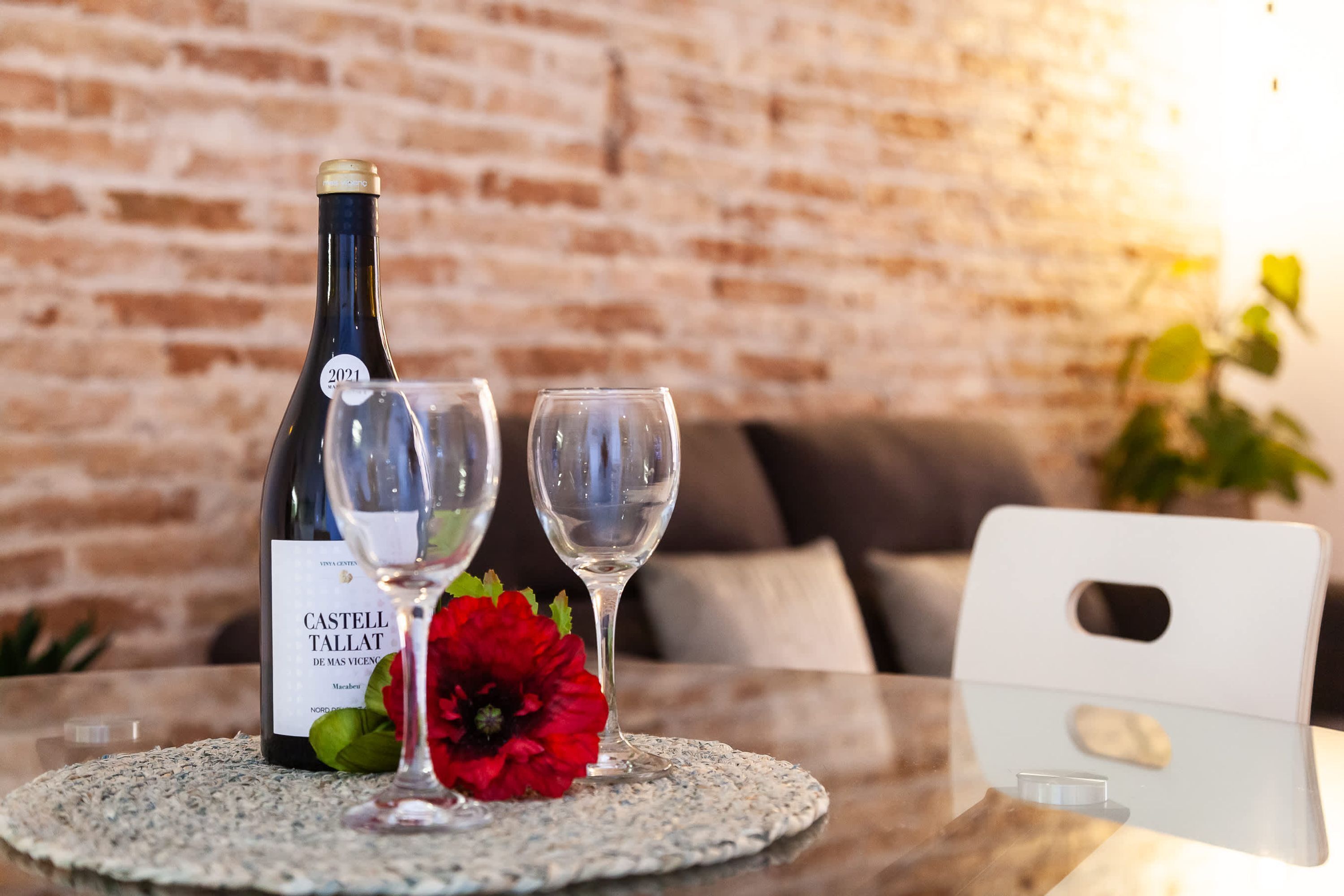 Une bouteille de vin de Castell Tallat, deux verres et une fleur rouge sur une table, avec un mur de briques en arrière-plan.