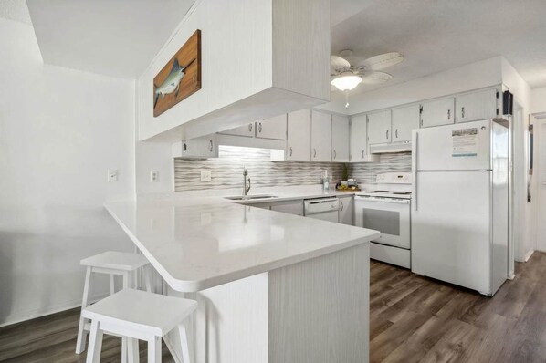Una cocina moderna con armarios blancos, encimera blanca y horno empotrado. Hay dos taburetes blancos y un ventilador de techo.