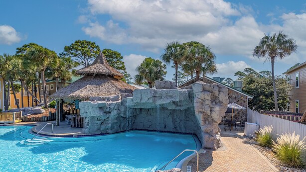 A swimming pool with a thatched-roof structure, a stone rock wall, and a paved patio area with outdoor seating.