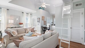 A spacious living room with a large beige sectional sofa, a dining table with chairs, and a white ladder against a wall.