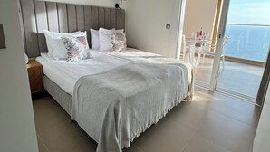 Ein modernes Schlafzimmer mit einem großen Bett, einem grauen Kopfteil und einem Balkon mit Meerblick.