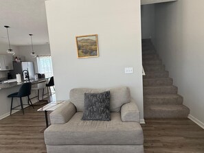 Un salon avec un canapé, un tableau au mur, un coin cuisine avec des tabourets de bar et un escalier.