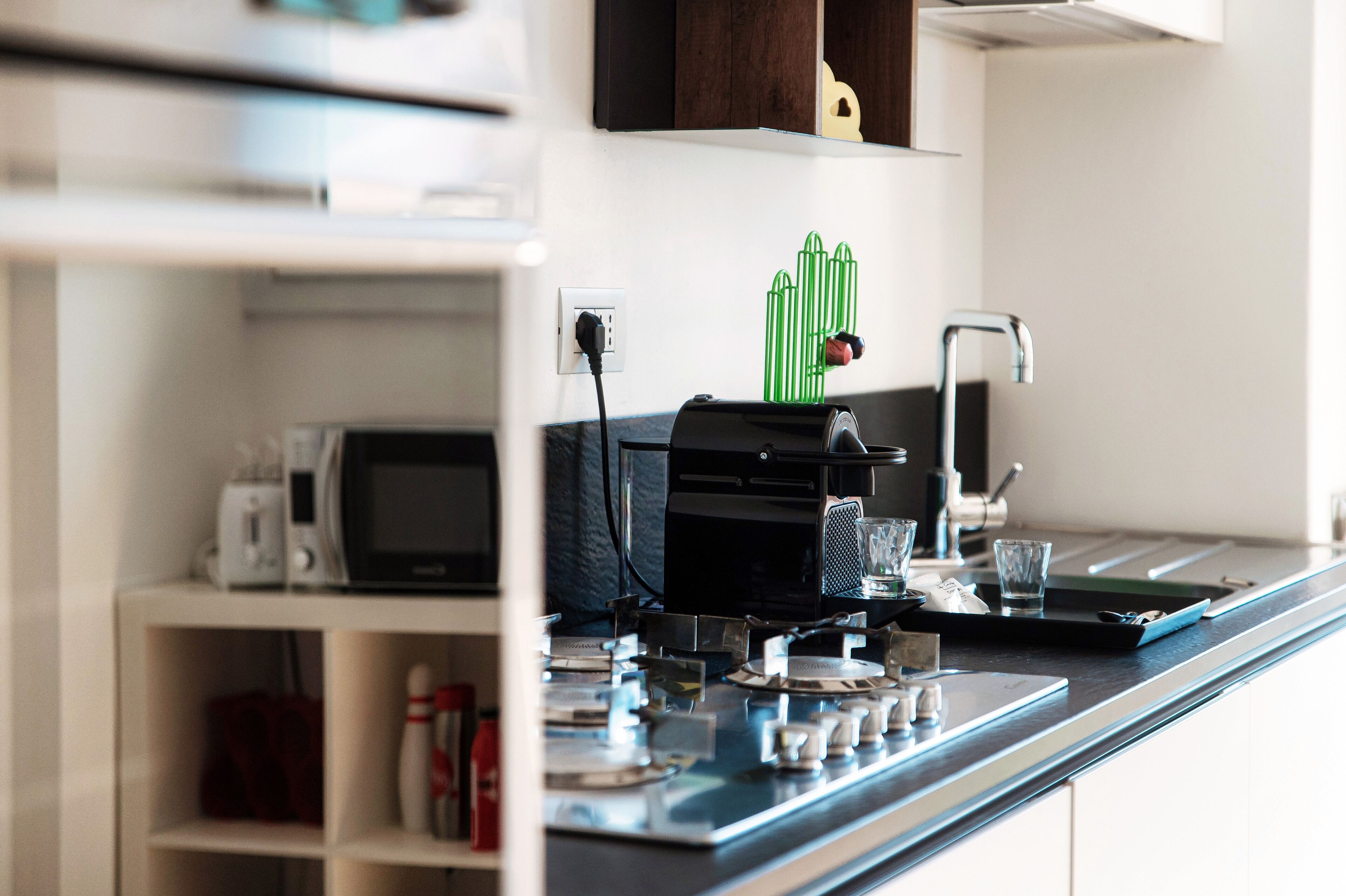 Un plan de travail de cuisine avec une machine à café, un four à micro-ondes et un grille-pain. Un objet décoratif vert est accroché au mur et une bouteille de sauce est posée sur le plan de travail.