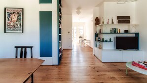 Una sala de estar moderna con piso de madera, una mesa de comedor, una estantería, un televisor y una cocina al fondo.