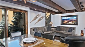 Una sala moderna con una mesa de comedor de cristal, sillas de cuero, un sofá gris y un ventanal con vistas a un paisaje nevado.