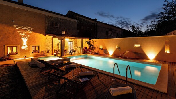 Ein Pool mit Liegestühlen und einer Holzterrasse, umgeben von einem Gebäude mit Steinwänden und warmer Beleuchtung.