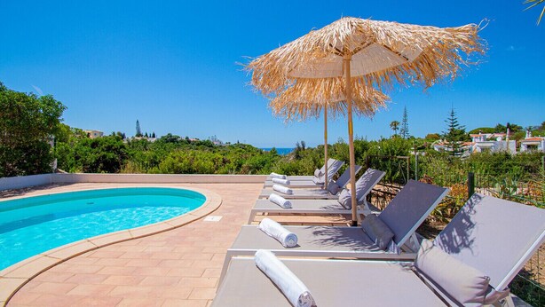 A poolside area with lounge chairs and a thatched umbrella, overlooking a clear blue sky and a distant coastline. - 91A Casa Mel: Stylish 3-Bed Townhouse with Pool (Carvoeiro)