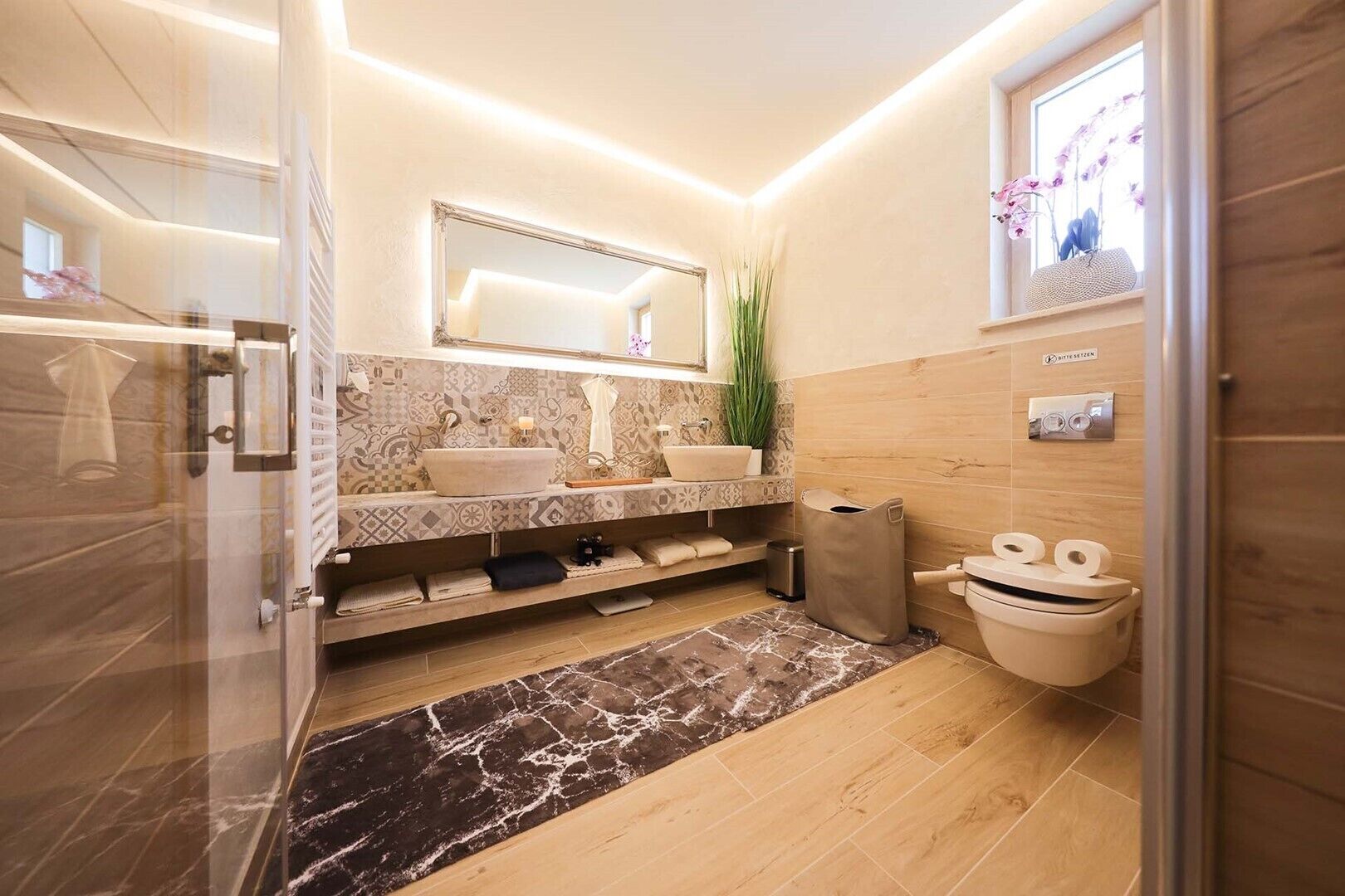 Un baño moderno con cabina de ducha de vidrio, tocador doble, inodoro y pequeña ventana con flores.