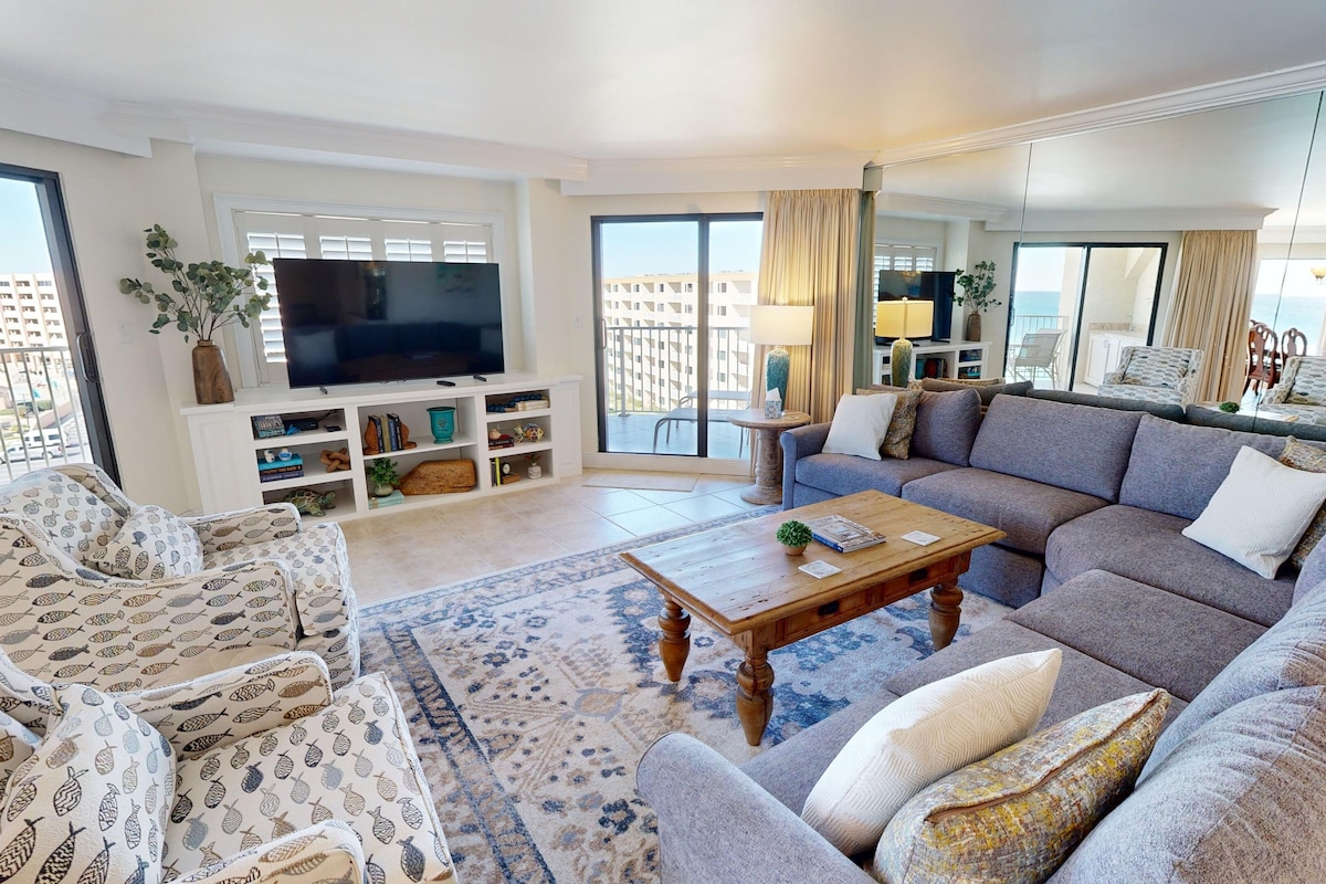 A spacious living room with a large sofa set, a coffee table, and a flat-screen TV. There is a balcony with a view of the city.