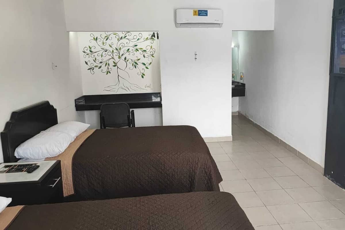 A hotel room with two beds, a desk, a chair, a wall-mounted air conditioner, and a TV displaying various channels.
